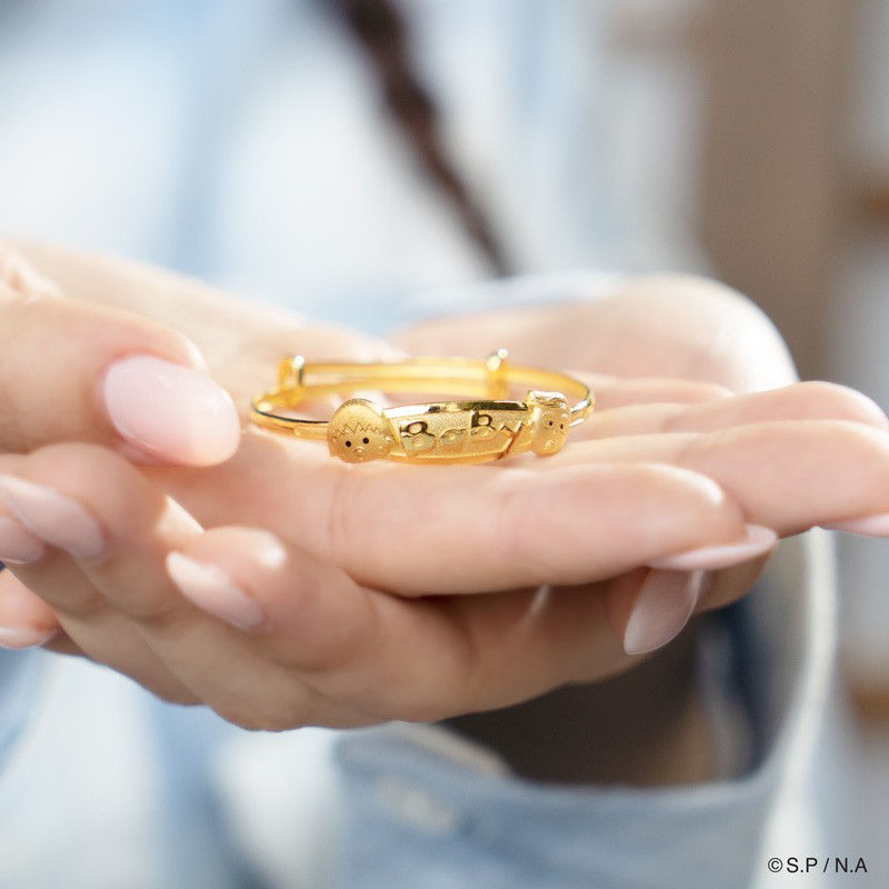 A hand holding a gold bracelet engraved with "Baby," symbolizing blessings for the baby's 100-day celebration.